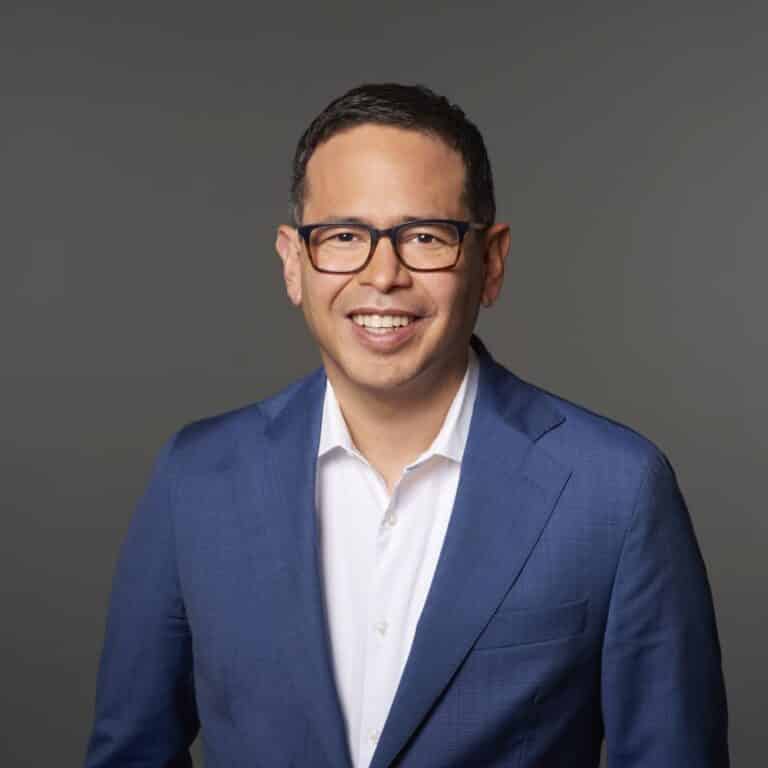 Mann mit Brille in blauem Anzug und weißem Hemd, lächelt vor grauem Hintergrund.