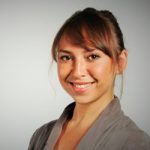 Jasmin Singgih Porträt einer lächelnden Frau mit braunen Haaren, die einen grauen Blazer trägt, vor einem hellen Hintergrund.