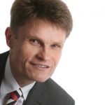 Stefan Schneider Porträt eines Mannes mit kurzem, braunem Haar, in einem Anzug mit Krawatte, lächelnd vor hellem Hintergrund.