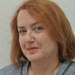 Caroline Rudolph Porträt einer Frau mit roten Haaren, die in die Kamera lächelt, vor einer hellen, strukturierten Wand.