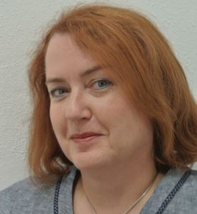 Caroline Rudolph Porträt einer Frau mit roten Haaren, die in die Kamera lächelt, vor einer hellen, strukturierten Wand.