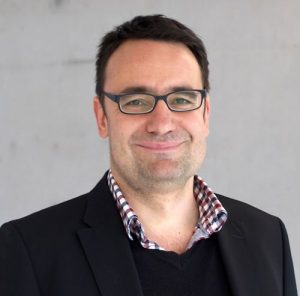 Mann mit Brille und dunklem Anzug, lächelt vor neutralem Hintergrund.