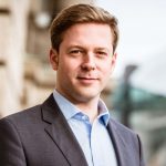 Helge Fuhst Porträt eines Mannes in Anzug mit kurzem, blondem Haar, stehend vor einem modernen Gebäude.