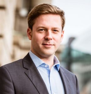 Porträt eines Mannes in Anzug mit kurzem, blondem Haar, stehend vor einem modernen Gebäude.