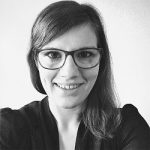 Porträt einer lächelnden Frau mit Brille, dunklem Oberteil und langen, braunen Haaren vor einem hellen Hintergrund.