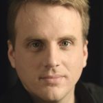 Kai Schlieter Porträt eines Mannes mit kurzen, blonden Haaren und grünen Augen, vor einem dunklen Hintergrund.