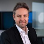 André Mücke Porträt eines Mannes mit grauen Haaren, in Anzug und Hemd, lächelnd vor einem modernen Hintergrund.