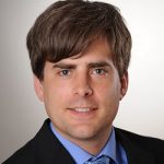Mathias König Porträt eines Mannes mit braunen Haaren, in Anzug und Krawatte, lächelnd vor einem hellen Hintergrund.