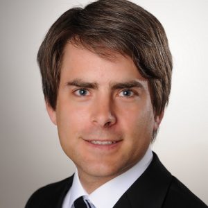 Wolfgang König Porträt eines Mannes mit braunen Haaren und blauen Augen, der einen schwarzen Anzug und eine Krawatte trägt.