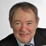 Ekkehard Seegers Porträt eines lächelnden Mannes mit Brille, grauen Haaren und schwarzem Anzug, vor hellem Hintergrund.