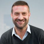 Lächelnder Mann mit kurzen Haaren und Bart, trägt einen grauen Pullover über einem hellblauen Hemd.