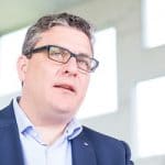 Mann mit Brille und Anzug spricht, Hintergrund mit großen Fenstern.