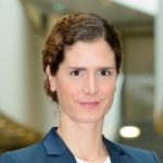 Porträt einer Frau mit lockigem Haar, in einem blauen Blazer, vor einem unscharfen, hellen Hintergrund.