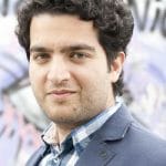 Porträt eines jungen Mannes mit lockigem Haar, der einen blauen Blazer und ein kariertes Hemd trägt, vor einem bunten Hi...