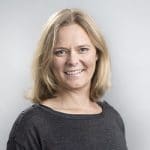 Porträt einer lächelnden Frau mit blonden Haaren, die in einem grauen Oberteil vor einem hellen Hintergrund steht.