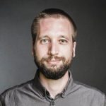 Portrait eines Mannes mit Bart, der in die Kamera lächelt, vor einem dunklen Hintergrund.