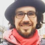 Mann mit Brille, Hut und rotem Schal lächelt in die Kamera, Hintergrund unscharf.