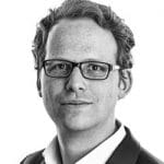 Porträt eines Mannes mit Brille, kurzen, lockigen Haaren und einem neutralen Gesichtsausdruck, vor hellem Hintergrund.
