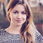 Porträt einer lächelnden Frau mit langen, blonden Haaren und blauen Augen, die ein gemustertes Oberteil trägt.