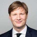 Porträt eines lächelnden Mannes mit blonden Haaren, in einem dunklen Anzug und weißem Hemd, vor einem grauen Hintergrund.