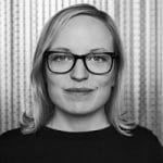 Porträt einer lächelnden Frau mit Brille, blonden Haaren und neutralem Hintergrund.