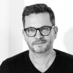 Porträt eines Mannes mit Brille, kurzen Haaren und Bart, lächelnd vor einem hellen Hintergrund.