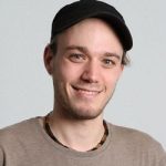 Porträt eines lächelnden Mannes mit kurzer Frisur, schwarzer Mütze und braunem T-Shirt, vor hellem Hintergrund.