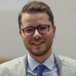 Porträt eines lächelnden Mannes mit Brille, hellgrauem Blazer und blauer Krawatte, vor unscharfem Hintergrund.