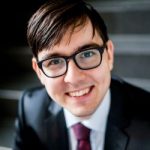Porträt eines lächelnden Mannes mit Brille, dunklem Anzug und Krawatte, vor einer Treppe.