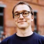 Mann mit Brille und kurzem Haar, lächelt vor einer roten Backsteinmauer.