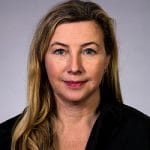 Porträt einer Frau mit langen, blonden Haaren, die in einem schwarzen Oberteil vor einem grauen Hintergrund steht.