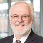 Burkhard Göke Porträt von Burkhard Göke, einem älteren Mann mit Brille, grauem Bart und Anzug, lächelnd vor einem unscharfen Hinterg...