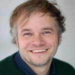 Tim Braune Lächelnder Mann mit kurzen, lockigen Haaren und blauen Augen, trägt einen grünen Pullover und ein dunkles Hemd.