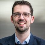 Dirk von Gehlen Mann mit Brille und dunklem Anzug lächelt vor einem unscharfen Hintergrund.