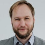 Neven Klepo Porträt eines Mannes mit Bart, lächelnd, in einem grauen Pullover und kariertem Hemd, vor neutralem Hintergrund.