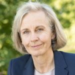 Ursula Münch Porträt einer Frau mit blonden, schulterlangen Haaren, in einem blauen Blazer, lächelnd vor unscharfem grünem Hintergrund.