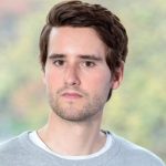 Bendix Hügelmann Porträt eines jungen Mannes mit braunen Haaren, neutralem Gesichtsausdruck, in einem grauen Sweatshirt vor unscharfem Hin...