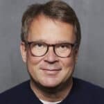 Porträt eines Mannes mit Brille und kurzem, graubraunem Haar vor einem grauen Hintergrund.