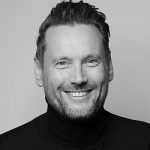Daniel Stich Porträt eines lächelnden Mannes mit kurzen Haaren, der einen schwarzen Rollkragenpullover trägt, vor neutralem Hintergr...
