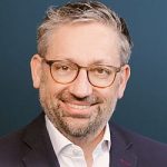 Heiko Kretschmer Porträt eines lächelnden Mannes mit Brille, grauen Haaren und einem dunklen Anzug vor einem blauen Hintergrund.