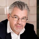 Stefan Krüger Porträt eines Mannes mit grauen Haaren, Brille und einem weißen Hemd unter einem dunklen Blazer, lächelnd in die Kamera...
