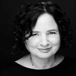 Beate Walter-Rosenheimer (Grüne) Porträt einer lächelnden Frau mit lockigem, schulterlangem Haar vor schwarzem Hintergrund.