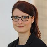 Porträt einer Frau mit langen, braunen Haaren, Brille und schwarzem Blazer, vor einem hellen Hintergrund.
