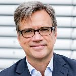 Frank Hartmann Mann mit Brille und grauen Haaren lächelt in Anzug vor einem hellen, gestreiften Hintergrund.
