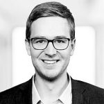 Sebastian Steinbach Porträt eines lächelnden Mannes mit Brille, in Anzug und Hemd, vor einem unscharfen Hintergrund.