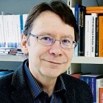 Uwe Jun Porträt eines Mannes mit Brille, lächelnd vor einem Bücherregal mit verschiedenen Büchern.