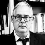 Uwe Wagschal Schwarz-weiß Porträt eines Mannes mit Brille, Anzug und Krawatte, vor einem Bücherregal.