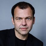 Marc Hujer Porträt eines Mannes mit kurzen, dunklen Haaren und blauen Augen, der einen schwarzen Blazer trägt. Hintergrund in Grau.