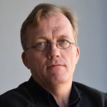 Markus Decker Porträt eines Mannes mit Brille, kurzen Haaren und einem nachdenklichen Ausdruck, vor einem hellen Hintergrund.