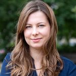 Porträt einer lächelnden Frau mit langen, lockigen Haaren, vor unscharfem grünem Hintergrund.